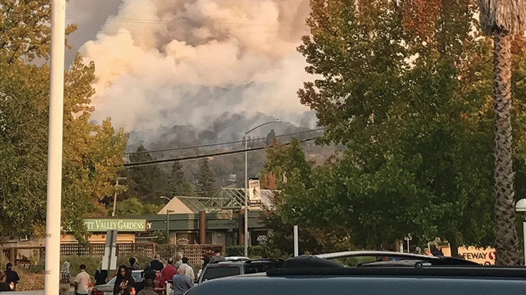 Smoke from a national wildfire bellows in the distance as people stand and look at it.