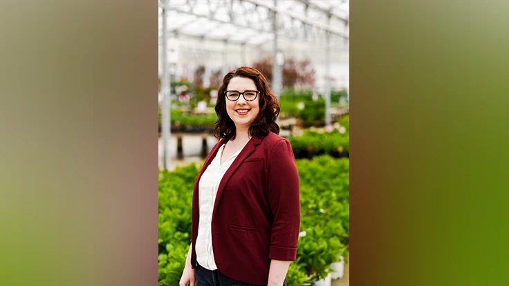 A smiling woman with shoulder-length reddish-brown hair wearing glasses, a maroon suit jacket and white shirt stands in a greenhouse surrounded by green plants.