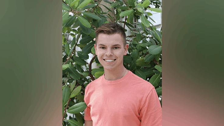 a man in a salmon colored t shirt in front of greenery