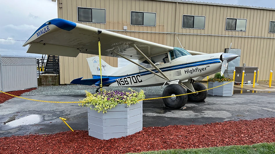 A white and red plane sitting on pavement in front of a building and behind a yellow chain rope and gray planters of flowers.