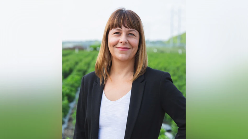 A woman in a black blazer and white shirt with shoulder-length brown hair and bangs smiles at the camera. The background shows rows of green plants in a field.