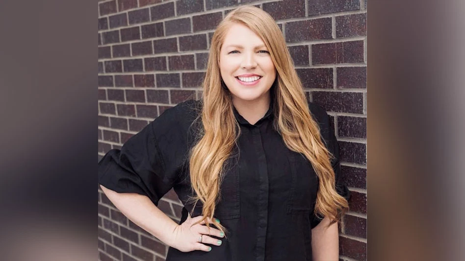 A smiling woman in a black shirt with long reddish blonde hair stands in front of a black wall with one hand on her hip.