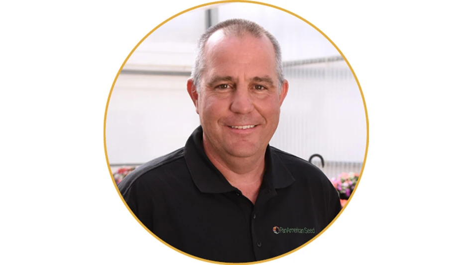 A smiling man with short gray hair wearing a black polo shirt stands in a greenhouse.