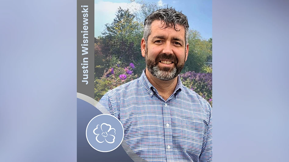 A smiling man with brown and gray hair and beard wearing a blue plaid shirt. He stands in front of lots of flowers, and text on the photo reads Just Wisniewski, with a graphic of a white outline of a flower on a pale blue background.
