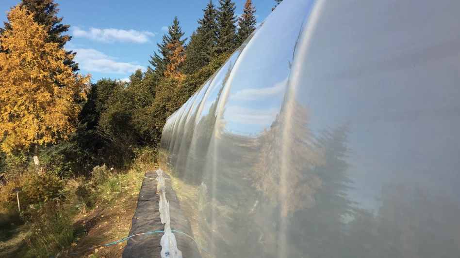 A side view of a poly greenhouses covering, with green, yellow and orange trees in the fall in the background.