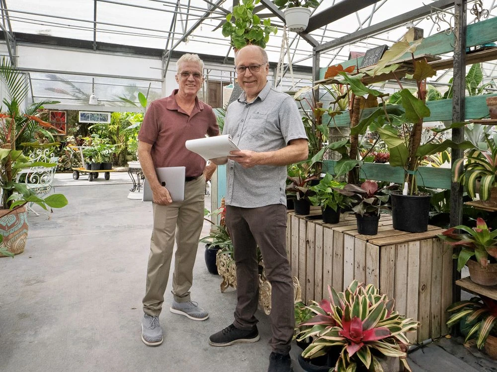Two smiling man stand in a retail greenhouse surrounded by plants.