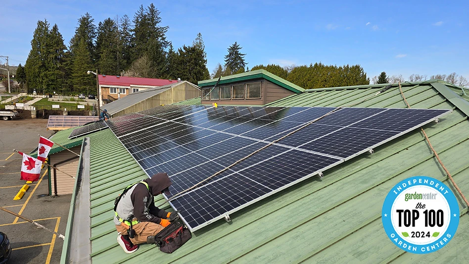 A person installs black solar panels on the green roof of a building. Tall green pine trees and a black parking lot are visible in the background.