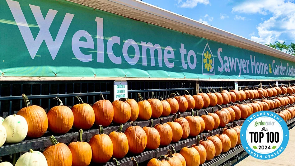 Three rows of orange pumpkins on black racks sit under a banner sign with a green background and white text that reads Welcome to Sawyer Home & Garden Center.