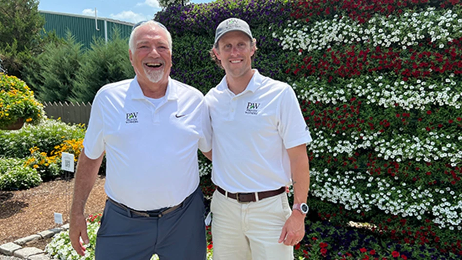 Two smiling men, both waring white polo shirts with a black PW logo on the chest, stand with their arms around each other in front of a flower wall made to look like the American flag. The man on the left has short white hair and a short white beard and wears blue pants. The man on the right has a gray baseball cap and wears khaki pants.