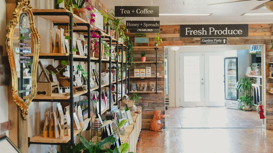 A store with wooden walls and tile floors, with wooden shelves with black piping holding a variety of products. Signs hanging from the ceiling above the shelves read Tea + Coffee, Honey + Spreads and Snacks. Additional signs above a wide doorway leading outside read Fresh Produce and Garden Party.