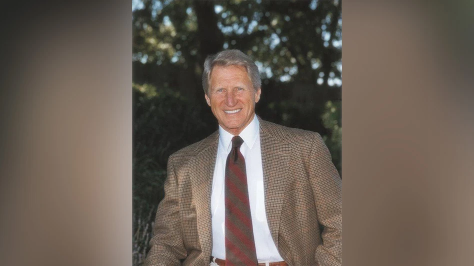 A smiling man with short white hair wearing a white shirt, brown and red plaid suit jacket and red and brown striped tie and standing in front of green trees.