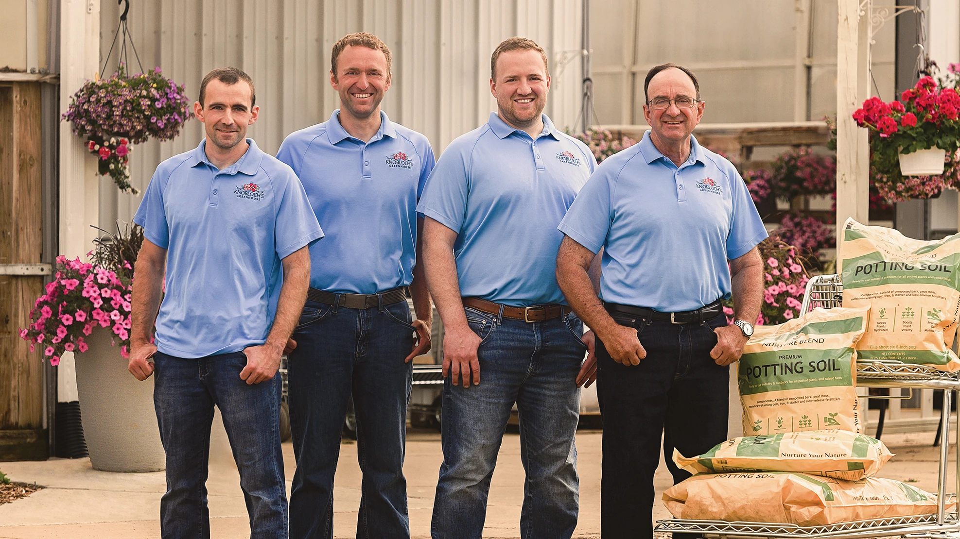 A photo of four smiling men wearing matching light blue polo shirts pose outside of a greenhouse.