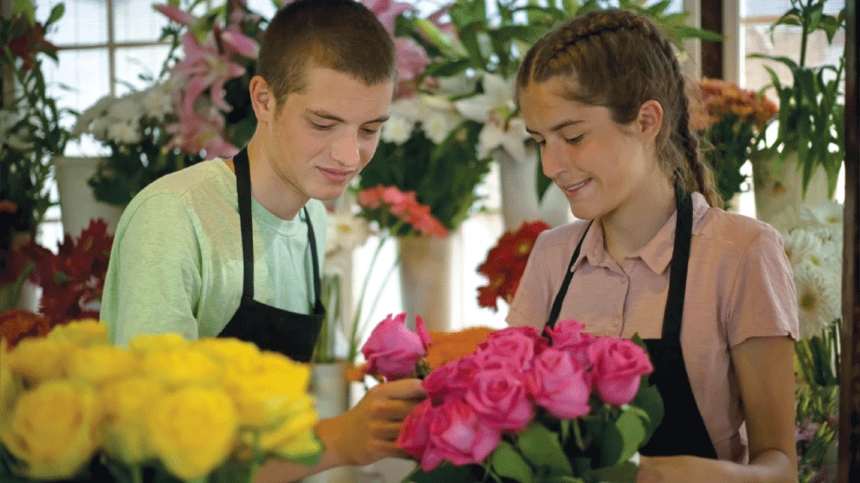 A teenage boy and teenage girl wearing black aprons stand in a shop surrounded by flowers. They're both reaching for a pink rose.