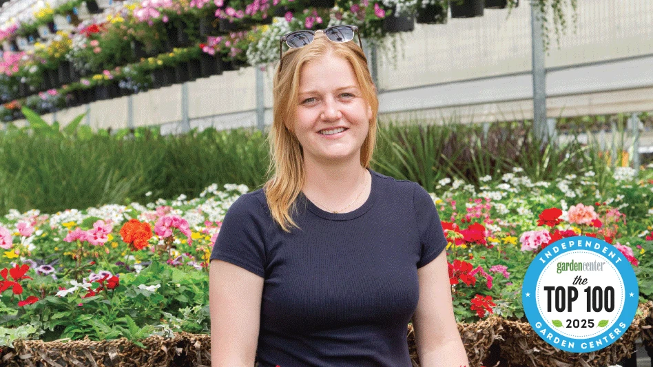 A smiling woman with shoulder-length blonde hair wearing sunglasses on top of her head and a black T-shirt stands surrounded by plants and flowers in a greenhouse.