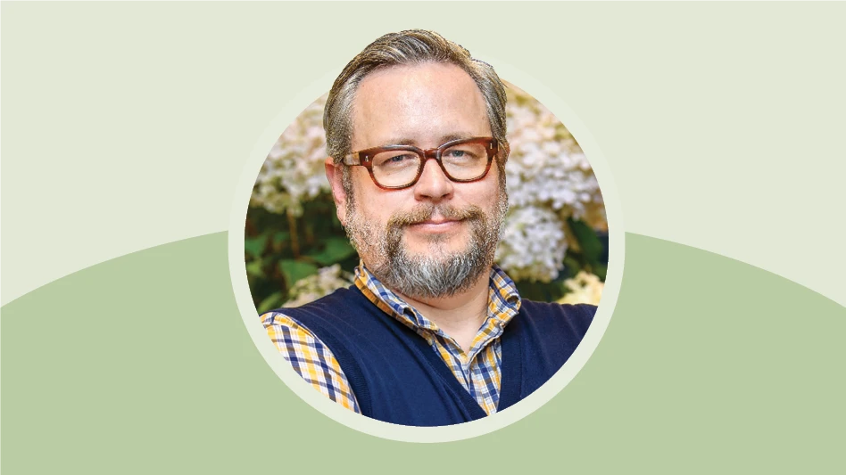 A man in a navy blue sweater vest with a white, yellow and blue plaid dress shirt underneath and wearing glasses crosses his arms and looks at the camera. There are flowers in the background behind him.