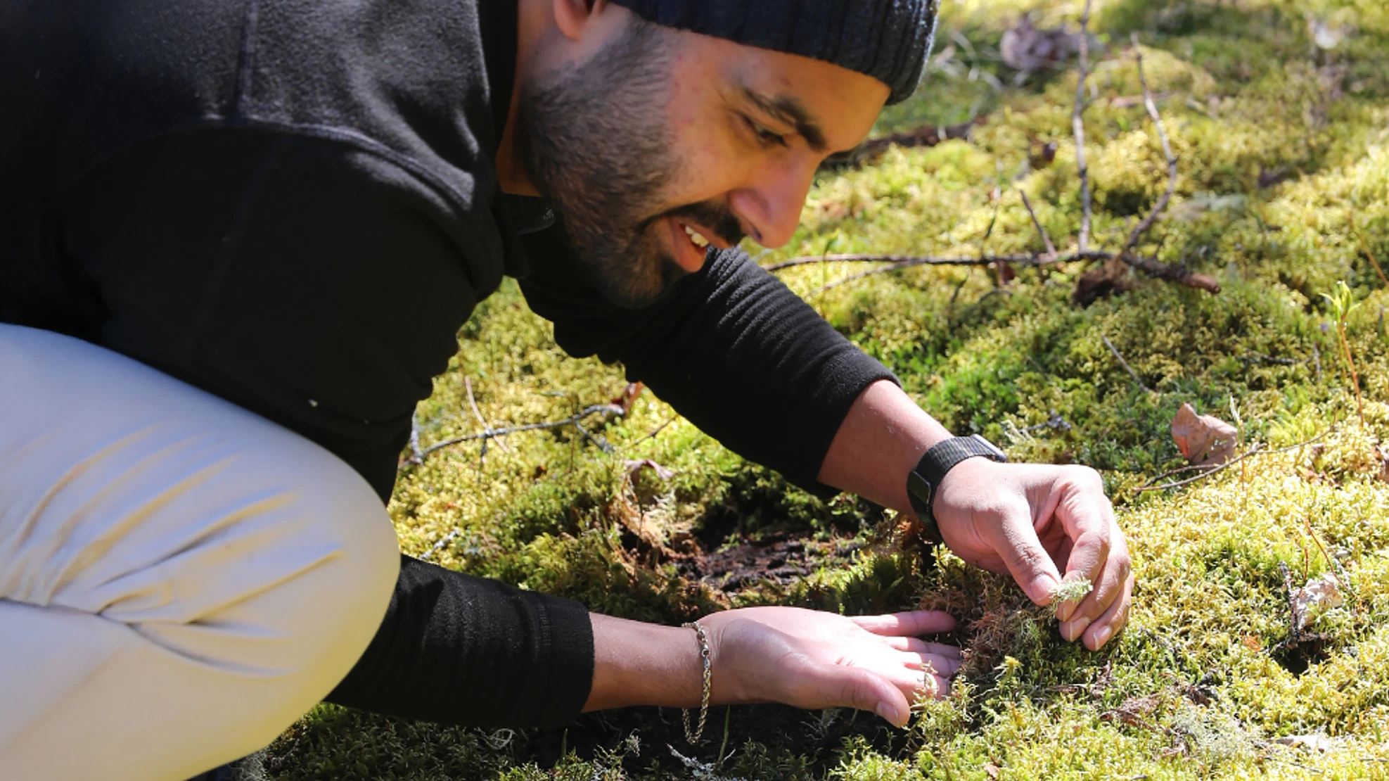 A man wearing a black hat, black shirt and khaki pants kneels down and looks at a small piece of moss he's holding in his hand.