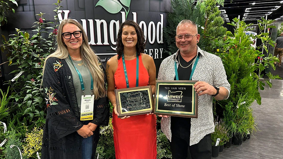 A man and two women stand in front of a wholesale nursery booth at a trade show. The man and one woman are holding a plaque award.