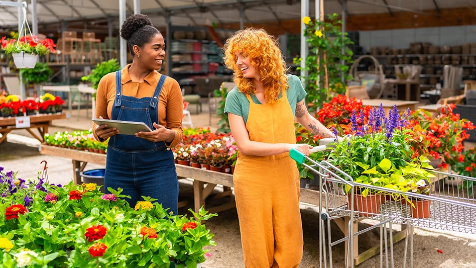 Two people looking at each other and smiling in a garden center surrounded by plants and flowers. The woman on the left is wearing an orange top and blue denim overalls and holding a tablet. The woman on the right is wearing a green top and orange overalls and pushing a silver metal cart with potted flowers.
