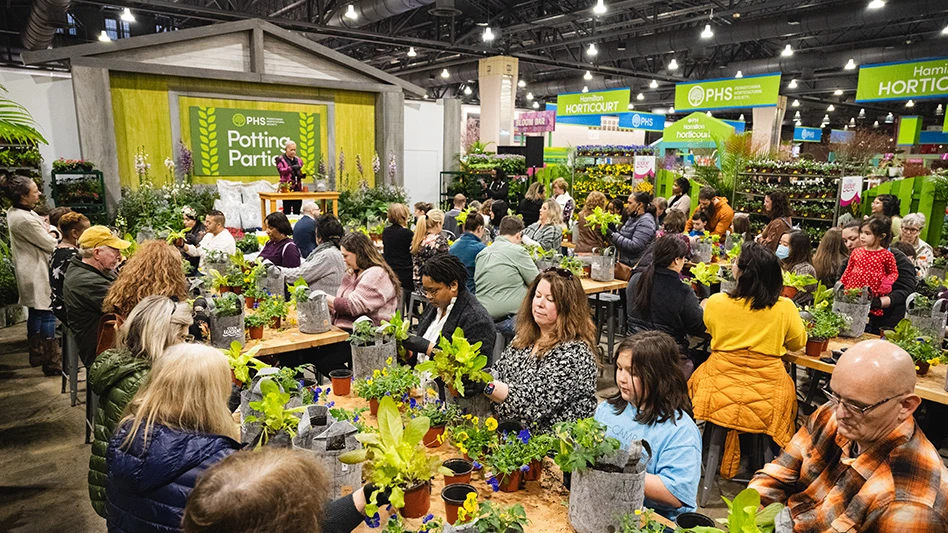 A workshop on a trade show floor, with several people sitting at wooden tables potting green plants in gray and orange pots.
