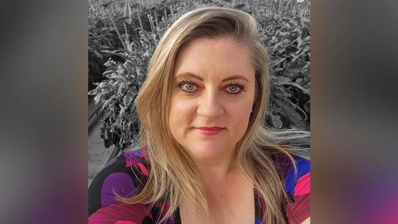 A woman with long blonde hair and hearing a black, purple, pink and blue shirt takes a selfie in front of gray plants.