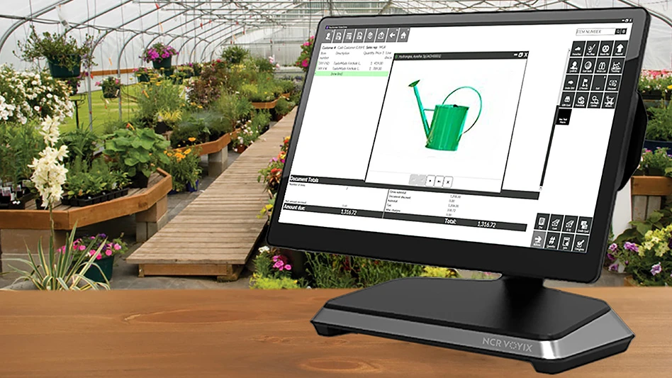 A black computer monitor showing a watering can on the purchasing screen sits on a brown wooden counter in a greenhouse, with potted plants and flowers and hanging baskets in the background.