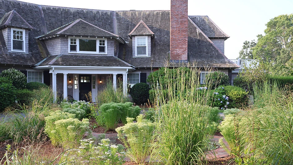 A large gray two-story house with a black roof and red chimney, with an elaborate garden in the front yard full of shrubs, grasses and plants.