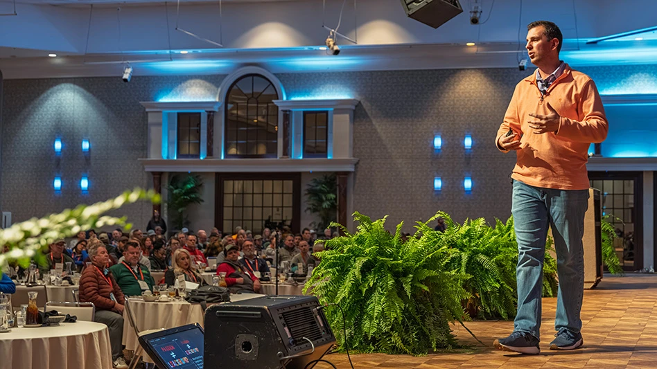 A man wearing jeans and an orange sweater addressing a crowd from a stage.