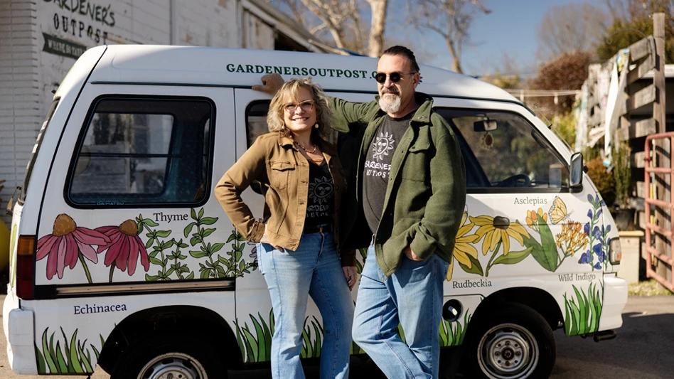 People sitting at a wooden picnic table in a gravel lot full of green plants and underneath red, blue and yellow shade awnings and hanging lights.