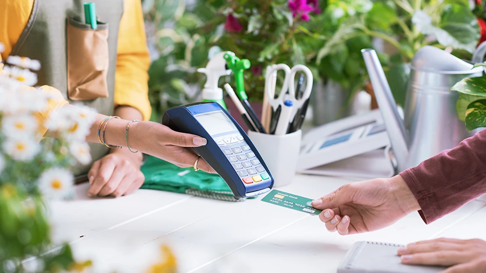 A person holds out a credit card payment machine while another person reaches toward it while holding a green credit card across a counter. The counter has potted plants on it.