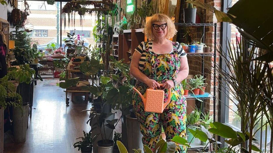 A smiling woman with chin-length blonde hair and wearing black glasses, a black and white striped shirt and a black apron with green leaves on it stands in a shop full of plants on wooden shelves.