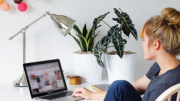 A woman in jeans and tshirt scrolls on laptop at a white desk.