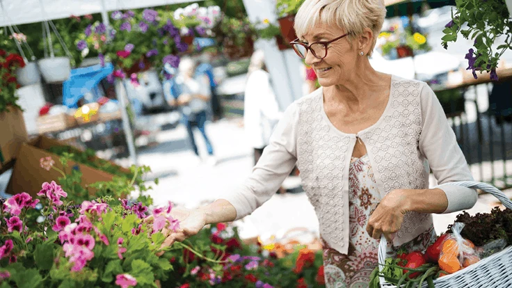 A woman in a beige cardigan and glasses is carrying a basket of plants and product. She is reaching for a display of pink flowers.